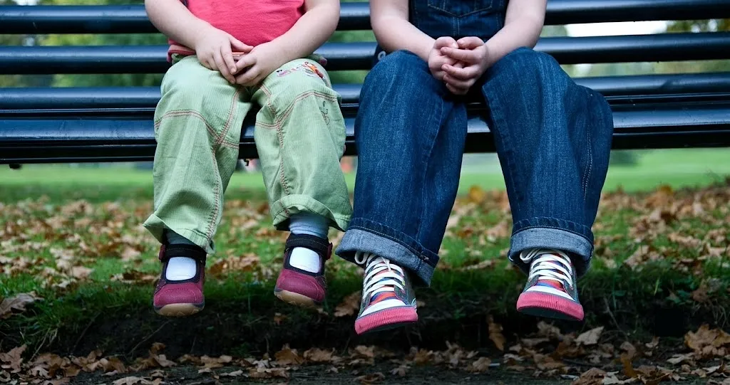 Two children sitting on a park bench, with the child on the left displaying feet pointing inward, illustrating the pediatric foot condition known as in-toeing or being pigeon-toed.