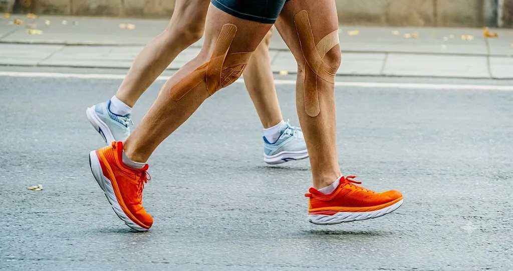 Close-up of a runner's legs wearing bright orange running shoes and kinesiology tape on the knee for support, highlighting common foot injuries in runners and how to avoid them.